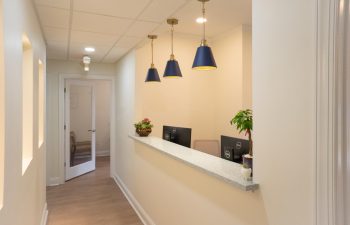 A hallway with beige walls and a counter desk equipped with two computer monitors. Above the desk hang three pendant lights with blue shades. A door at the end of the hallway is partially open.