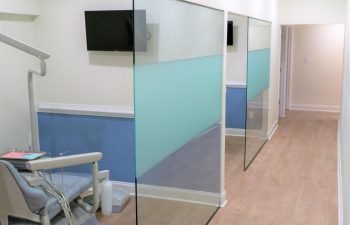 Dental office hallway with partitioned treatment areas featuring modern dental chairs and wall-mounted flat screen TVs. The flooring is light-colored wood, and the walls are white and blue.