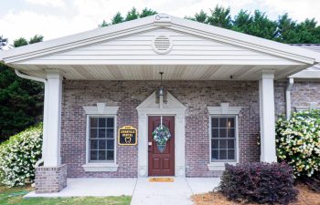 Front view of a brick building with a dark wooden door decorated with a wreath, flanked by two windows. A sign next to the door reads "Leahyla Beta II". A triangular pediment tops the entrance.