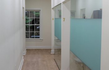 A clean hallway with light wooden floors, white walls, and numbered frosted glass cubicles on the right. A window is at the end of the hall, and a white chair is partially visible in cubicle 7.