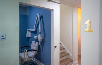 A small dental examination room with a blue accent wall, white dental chair, and equipment. The room is located on the right side of a hallway with stairs, indicated by a yellow number 1 on the wall.