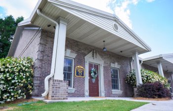 A single-story brick building with white trim has a wreath on the red door and a sign above it. There are flowering shrubs along the side and clouds in a blue sky overhead.