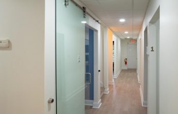 A hallway with a wooden floor features a sliding glass door to the left, an exit sign at the end, and numbered doors along the right wall.
