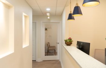 A modern lobby area with a reception desk, computer monitor, and decorative plants. Hallway with chair and glass door in the background. Hanging pendant lights illuminate the space.