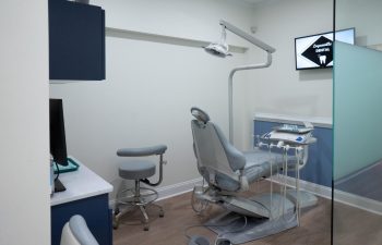 A modern dental office examination room featuring a dental chair, an overhead light, dental equipment, and a stool. A sign on the wall reads "Signature Dental.