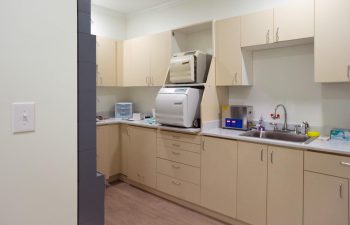 A clean, modern laboratory room with beige cabinetry, a large white machine, various small instruments, a sink, and storage compartments.
