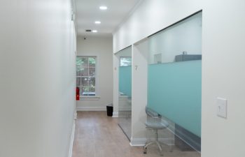 A hallway with white walls and wooden floors leads to office cubicles with frosted glass. A chair is in one cubicle, and natural light filters through a window at the end.