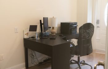 A home office with a black desk featuring a computer, monitor, and lamp. A black chair is positioned at the desk, and various electronic devices are arranged on and around the desk.
