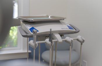 A dental workstation with various tools and instruments is positioned in a corner of an examination room, with a light blue chair and natural light from a window in the background.