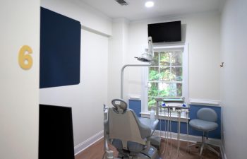 A modern dental examination room with a dental chair, equipment, and a wall-mounted TV. The room is well-lit with natural light from a window. Room number 6 is visible on the wall.