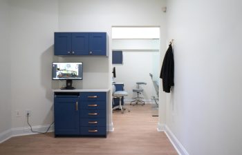 A clean room with blue cabinets, a desktop computer, and a coat rack. Another room with office chairs and more cabinetry is visible through an open doorway.