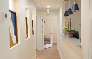 A clean hallway with light-colored walls, wooden floor, framed artwork, and blue pendant lights. A reception desk with flowers is on the right, and a glass door is at the end of the hall.