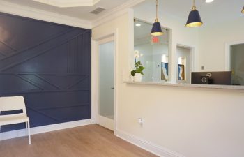A modern reception area with a white chair, dark blue accent wall, and a reception counter with hanging lamps and a potted plant.