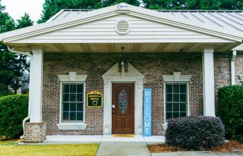 A brick dental office entrance with a brown door, a "Welcome" sign on the right, and a yellow "Loaraville Dental" sign on the left. The building is surrounded by greenery.