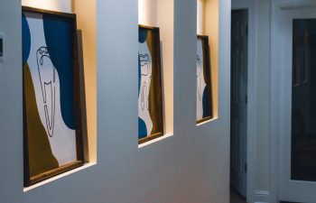 Three abstract art pieces in recessed wall niches, illuminated by downlights, line a hallway with wooden flooring.