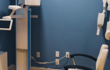 Dental X-ray machine in a blue-walled room, with an empty beige dental chair and various dental equipment visible.