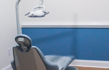 A dental operatory room with a reclining dental chair, overhead light, and a wall-mounted flat-screen TV. The room has blue and white walls and a wood-look floor.
