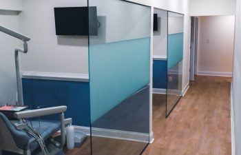 A dental office hallway with wooden flooring, frosted glass dividers, and an empty dental chair in an examination room. A wall-mounted TV is visible above the chair.