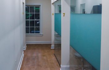 A hallway with wood flooring leads to numbered cubicles with frosted glass partitions, and a window at the end shows greenery outside.