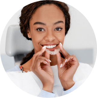 A woman sitting in a dental chair and holding a clear orthodontic aligner near her teeth, smiling.